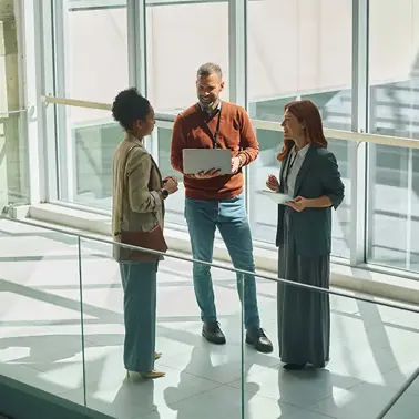 Three colleagues standing together in an office building, one holding an open laptop.