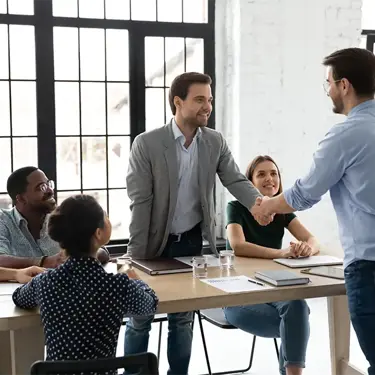 Two colleagues shaking hands in a meeting room, with another colleague in the background.