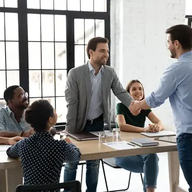 Two colleagues shaking hands in a meeting room, with another colleague in the background.