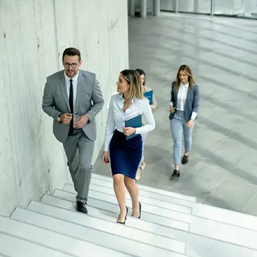 Two colleagues walking up the stairs in an office building while talking, with two other colleagues in the background.