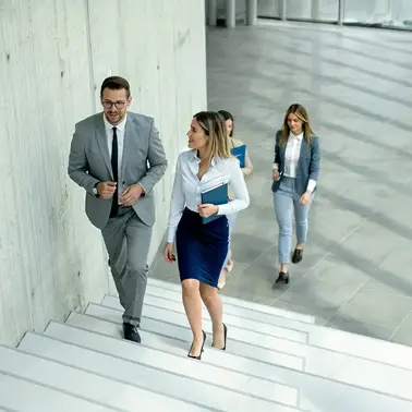 Two colleagues walking up the stairs in an office building while talking, with two other colleagues in the background.